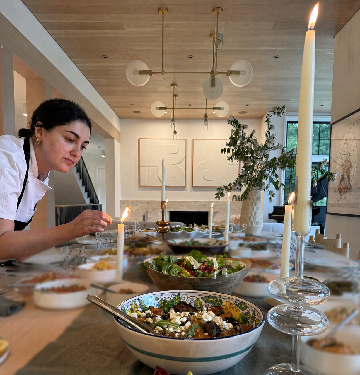 Chef in white chef coat lights candles at a catered table with salads, bowls, and warm chandelier lighting.